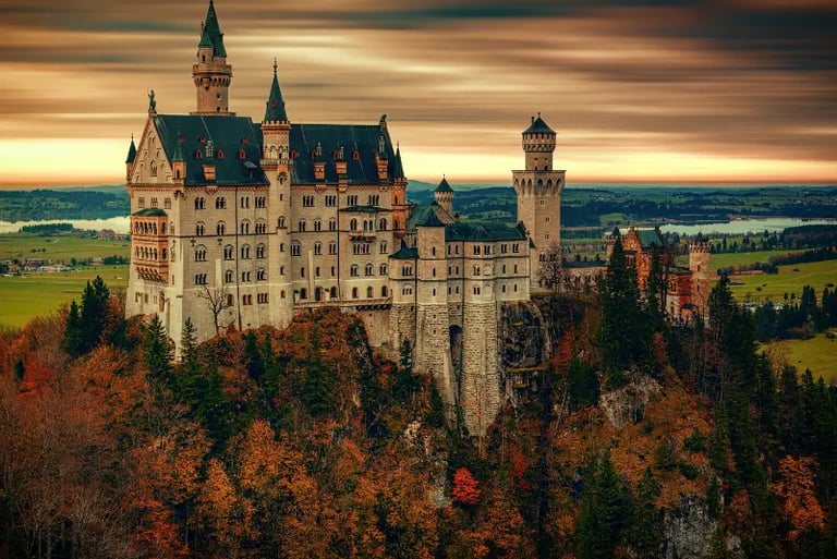 The Neuschwanstein Castle in Bavaria, Germany, stands atop a hill surrounded by autumn foliage at sunset.