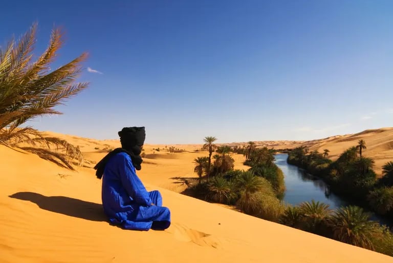 A man in traditional blue Tuareg clothing sitting on a Sahara Desert sand dune overlooking a lush river oasis.