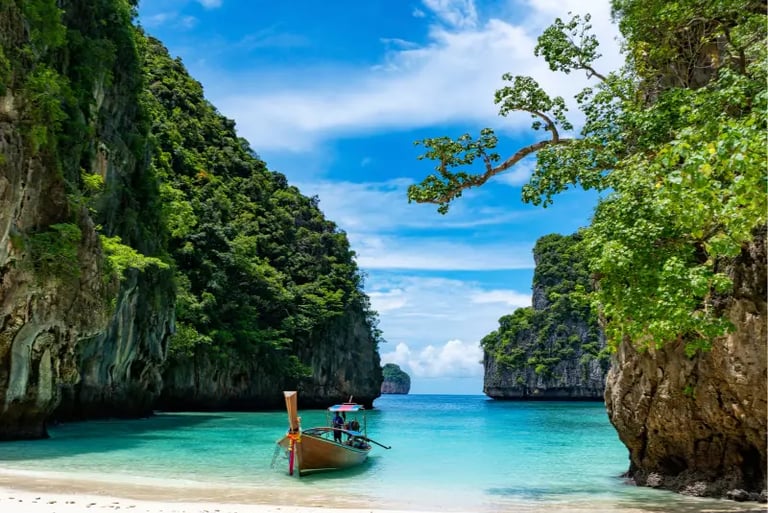 A traditional Thai longtail boat floats on turquoise water at a tropical beach surrounded by limestone cliffs.