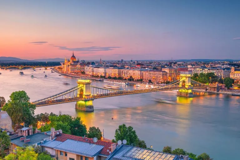 Panoramic sunset view of the illuminated Chain Bridge and Hungarian Parliament Building over the Danube River in Budapest.