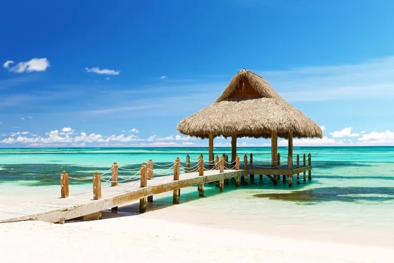 Tropical beach boardwalk leading to a thatched roof gazebo over turquoise ocean water under a blue sky.