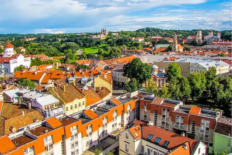 Panoramic view of Vilnius Old Town with orange terracotta rooftops, historic churches, and lush green hills.