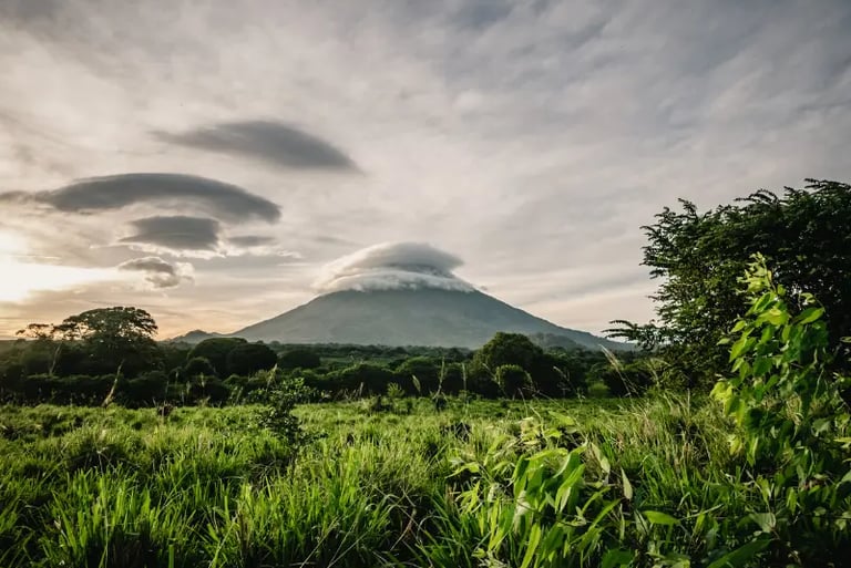 Scenic view of a tropical volcano peak topped with white lenticular clouds overlooking a green meadow.