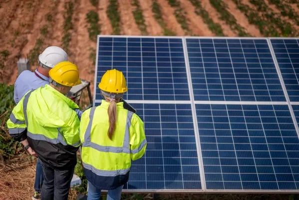 Fotografía de dos hombres y una mujer con cascos de protección color amarillo frente a paneles solar