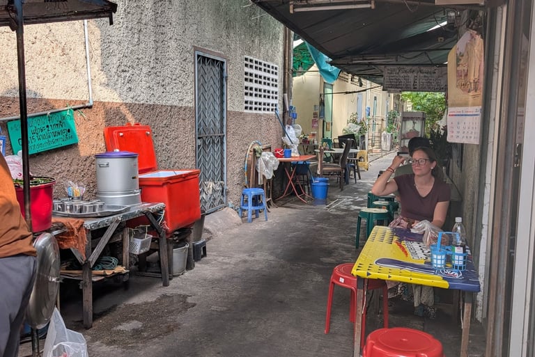 A local street food stall in a narrow Bangkok alley with plastic stools and outdoor seating.