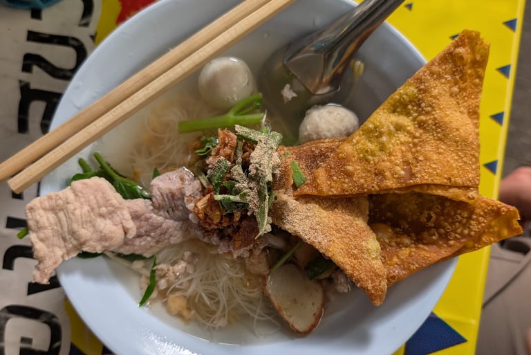 A bowl of Thai noodle soup with crispy wontons, fish balls, and pork on a yellow patterned table.