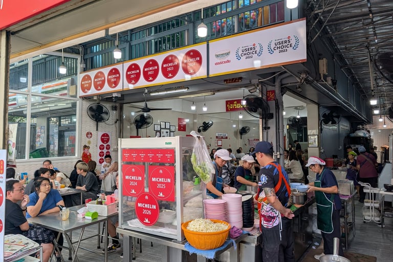 Diners eat at an open-air Michelin Guide street food stall in Bangkok featuring red award stickers.