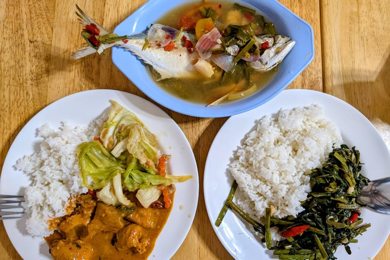 Thai steamed fish soup with white rice, red curry chicken, and stir-fried morning glory on a wooden table.