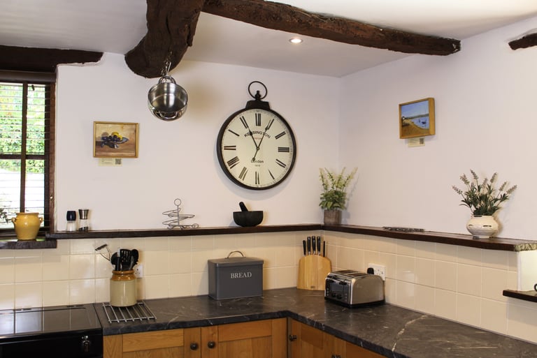 Rustic kitchen interior featuring exposed ceiling beams, a large wall clock, and grey marble countertops.