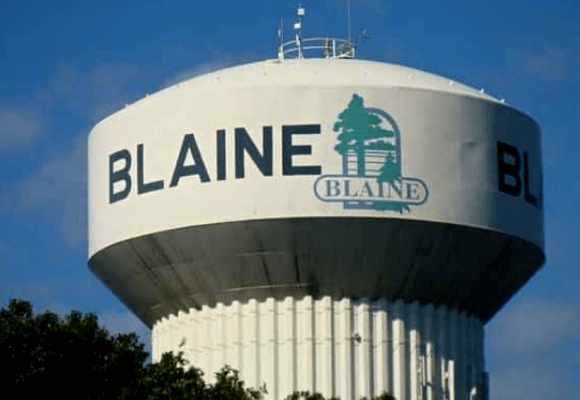 A large white municipal water tower in Blaine, Minnesota, featuring the city logo against a clear blue sky.