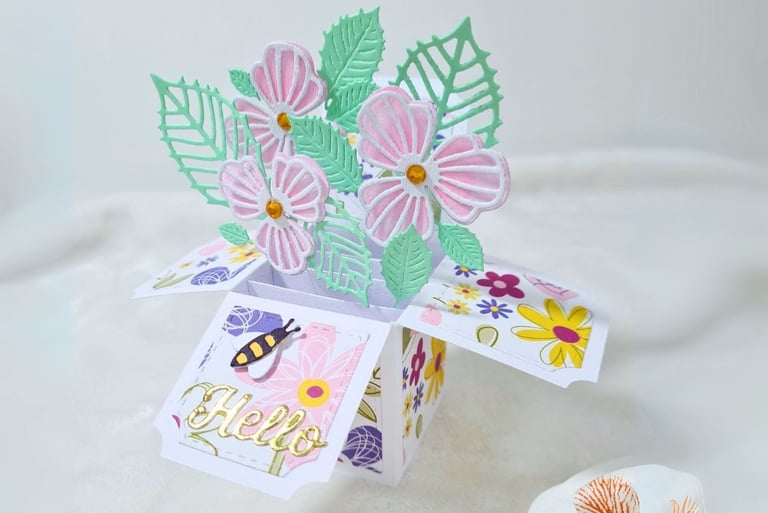 Pop-up card shaped like a box with pastel pink flowers and green leaves. "Hello" in gold script on a