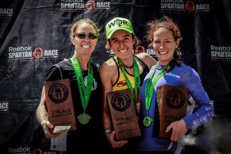 Three female athletes holding trophies on the Reebok Spartan Race podium after finishing the Beast Women's Elite.