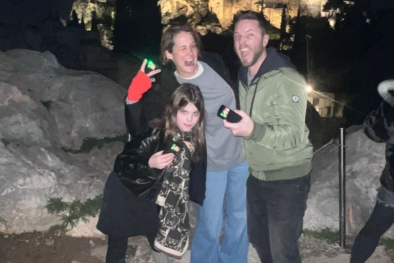 Family of three posing with green flashlights at night with illuminated Acropolis in background during Athens Ghost Tour