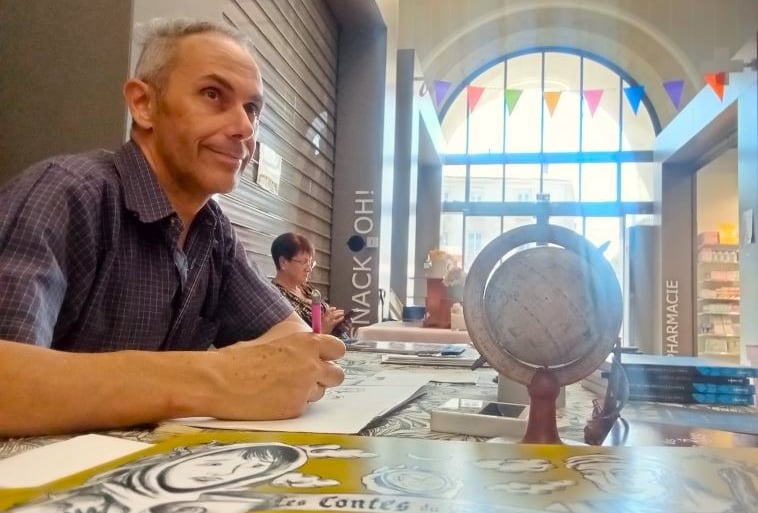 Artist Dim signing books at a table with a globe and colorful bunting in a bright indoor market.