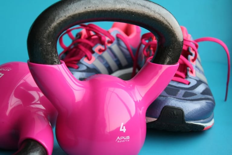 A pink kettlebell sits on a blue surface with tennis shoes in the background.
