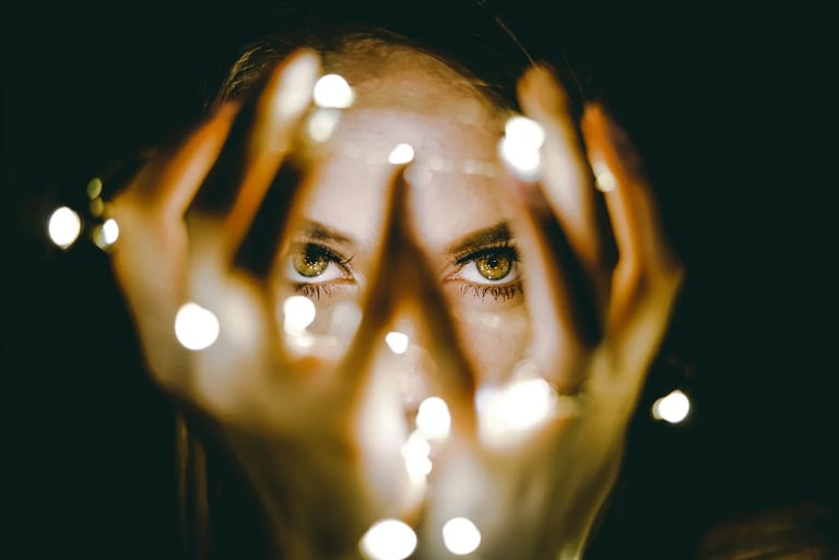 A woman with green eyes peeks through bokeh string lights held in her hands against a dark background.