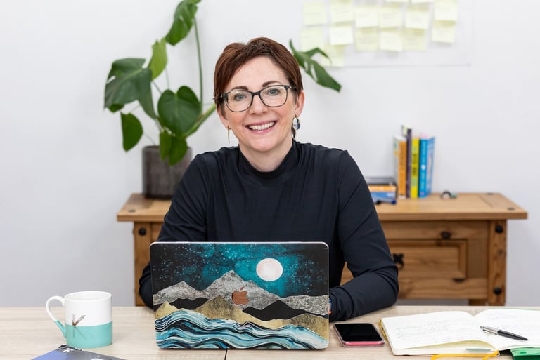 Kelly sat with her laptop in front of her and a large plant and some post-it notes in the background
