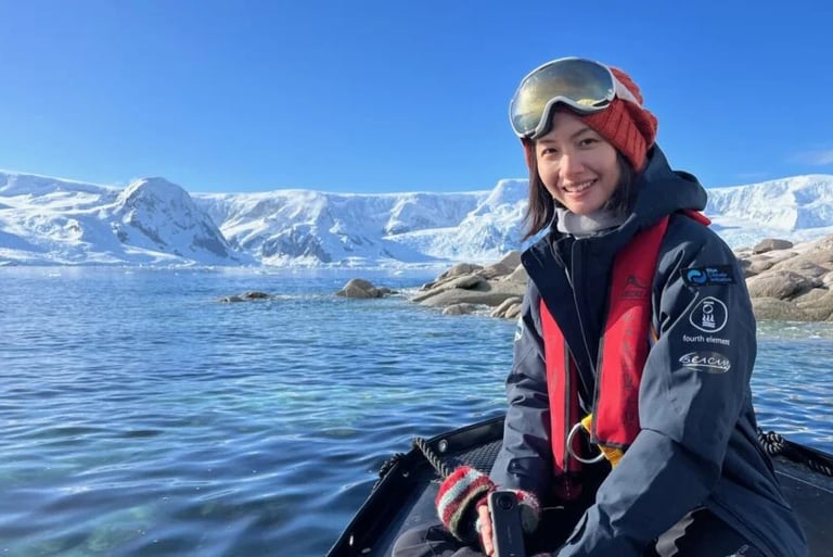 A smiling woman in polar gear on a boat in Antarctica with icy mountains in the background.