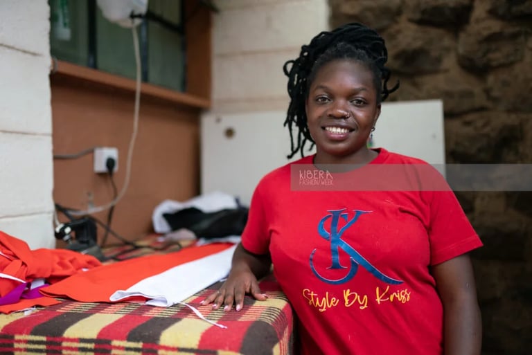 Smiling fashion designer at Kibera Fashion Week posing next to fabrics in a red Style By Kriss t-shirt.