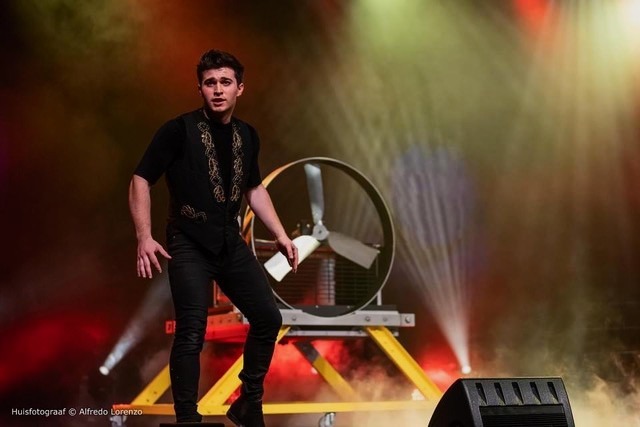 A male magician in a black vest performs on a stage with dramatic lighting and a large prop fan.