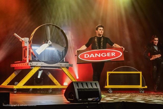 Magician Alfredo Lorenzo performing a dangerous stage illusion with a large industrial fan and a danger sign.