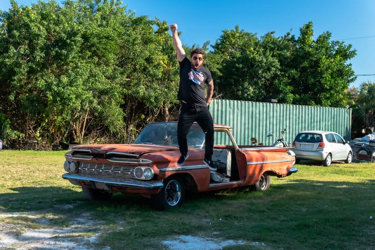 A man jumps with excitement on the hood of a classic rusty orange vintage car outdoors.