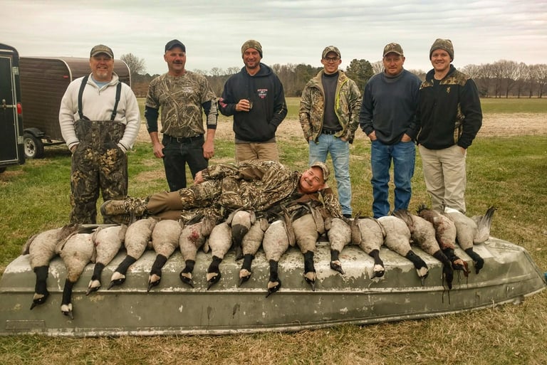 Five hunters standing behind a boat loaded with harvested Canada geese on a grassy shoreline.