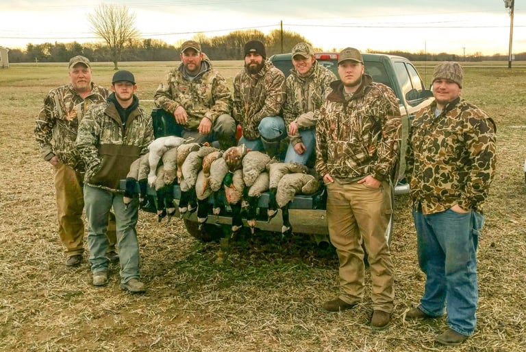 Six hunters in camouflage gear standing in a field behind a pickup truck loaded with harvested ducks and geese.