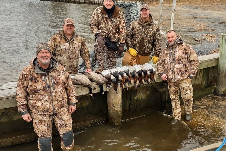 A hunting party posing on a dock in the water with a diverse harvest of ducks displayed in front of them.