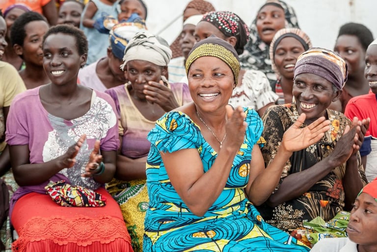 A group of smiling African women wearing colorful traditional clothing while clapping at a community gathering.
