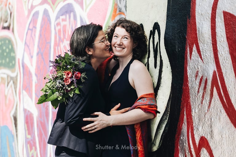 Two brides cuddling next to a wall with graffiti.