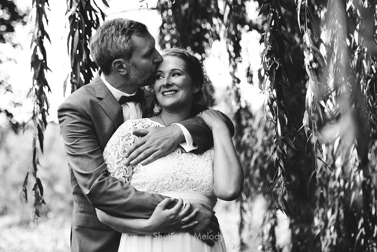 Groom hugging bride from behind a willow tree.
