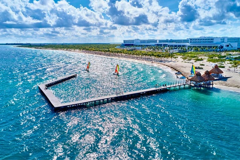 Aerial view of a luxury beachfront resort with a long wooden pier, turquoise water, and windsurfers.