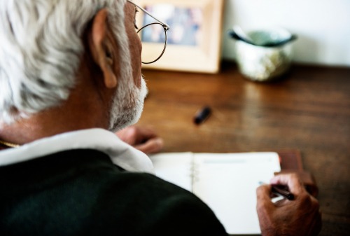 Senior man with white hair and glasses writing notes in a paper notebook at a wooden desk.
