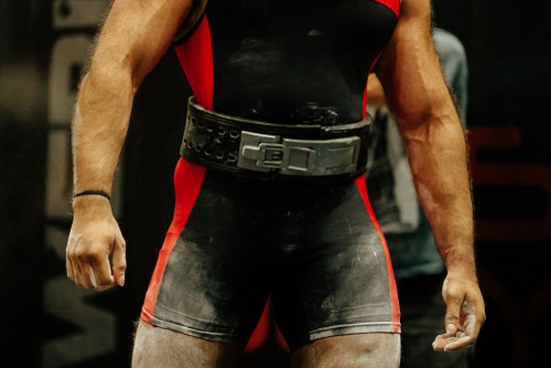 A muscular powerlifter wearing a red and black singlet and a heavy-duty lever lifting belt.