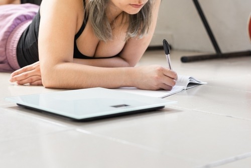 A woman lying on a floor writing in a weight loss journal next to a digital bathroom scale.