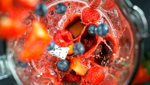 Fresh blueberries and raspberries splashing into a red berry fruit smoothie inside a glass blender.