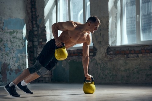 A shirtless muscular man performing a kettlebell renegade row in a rustic gym setting.