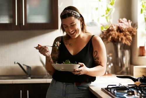 A smiling tattooed woman in a leopard headband enjoys a fresh green salad in a sunny kitchen.