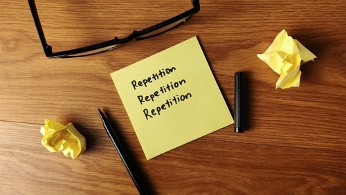 Yellow sticky note with the word repetition handwritten on a wooden desk with a pen and glasses.