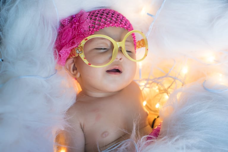 A newborn baby wearing oversized yellow glasses and a pink headband sleeps peacefully.