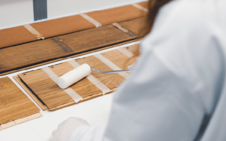 A person in a lab coat uses a small paint roller to apply a clear finish to wood floor samples.