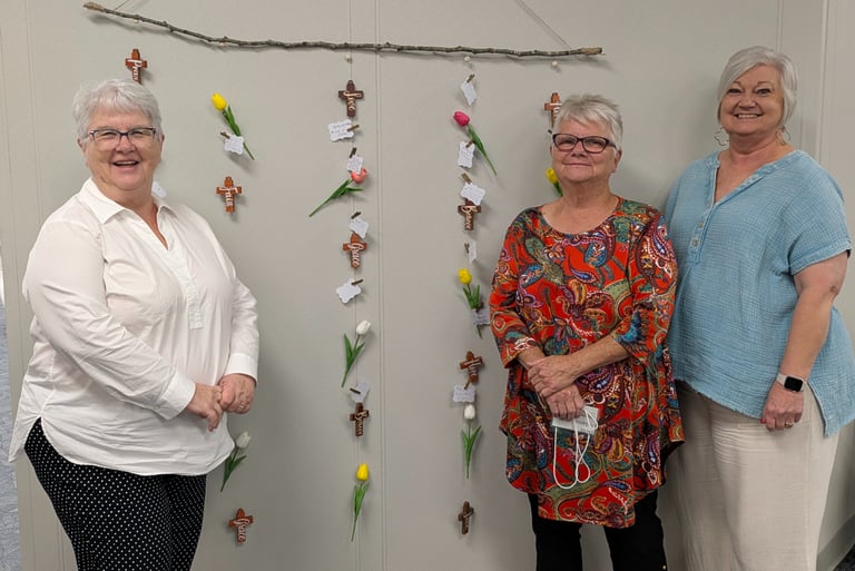 Three smiling women stand beside a hanging prayer wall display with wooden crosses and tulips.