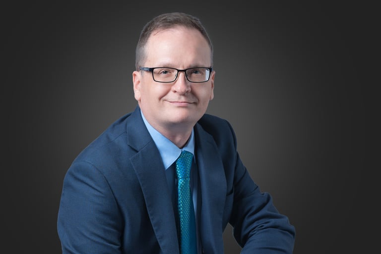 Professional corporate headshot of a smiling businessman wearing glasses, a blue suit, and a teal tie.
