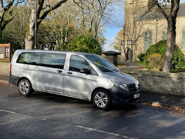 Silver Mercedes-Benz Vito wedding taxi parked outside a traditional stone church with autumn trees.