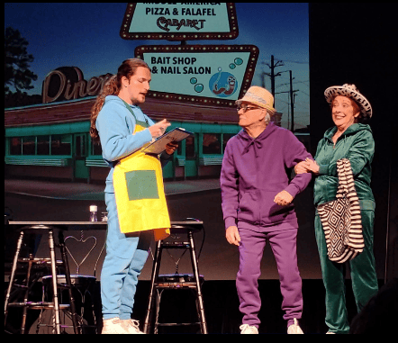 Actors in colorful tracksuits perform a comedic theater scene in front of a retro diner backdrop.