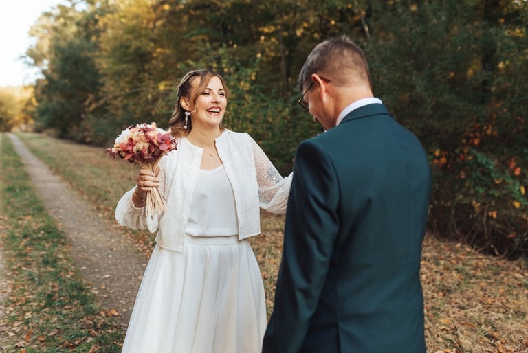 Le marié découvre la mariée et lui tend la main, tous deux souriants au cœur d'une allée automnale.