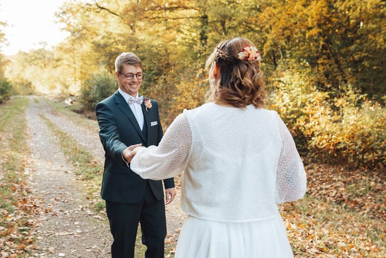 Le marié découvre la mariée et lui tend la main, tous deux souriants au cœur d'une allée automnale.