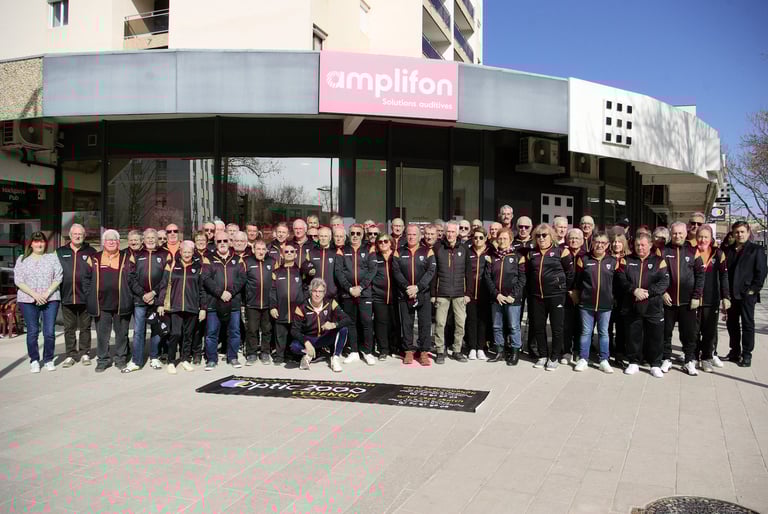 A large sports team in matching black and orange tracksuits posing outside an Amplifon storefront.