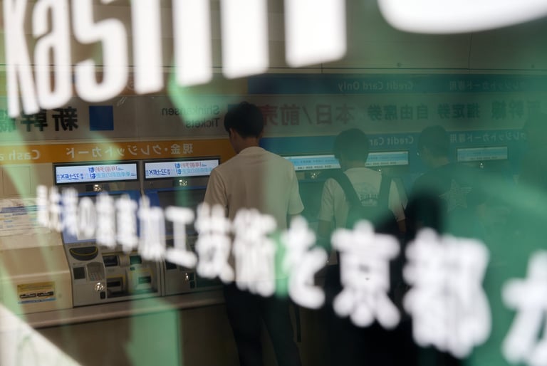 Commuters buying tickets at automated train station kiosks in Japan through a glass window.